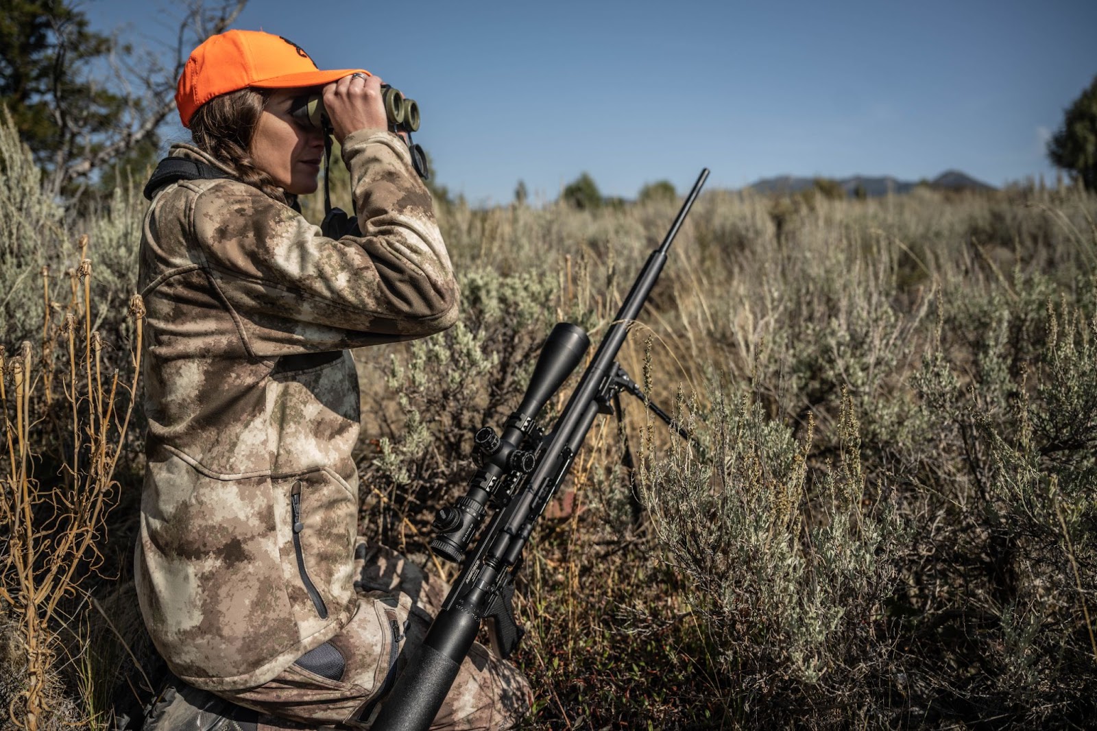 Air gun hunter looking through binoculars over the landscape for quarry while sitting next to their scoped rifle. 