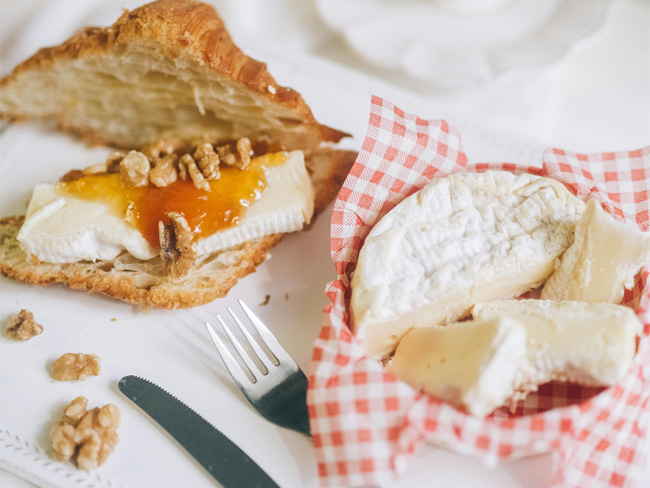 brie on a red and white checkered cloth, next to a piece of bread with a slice of brie, a dollop of marmalade, and some walnuts. 