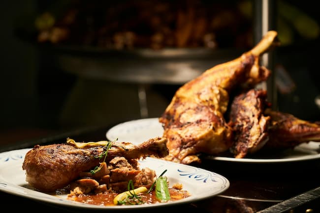 a dish of roasted rabbit leg and veggies next to a serving tray with the remainder of the roasted rabbit.